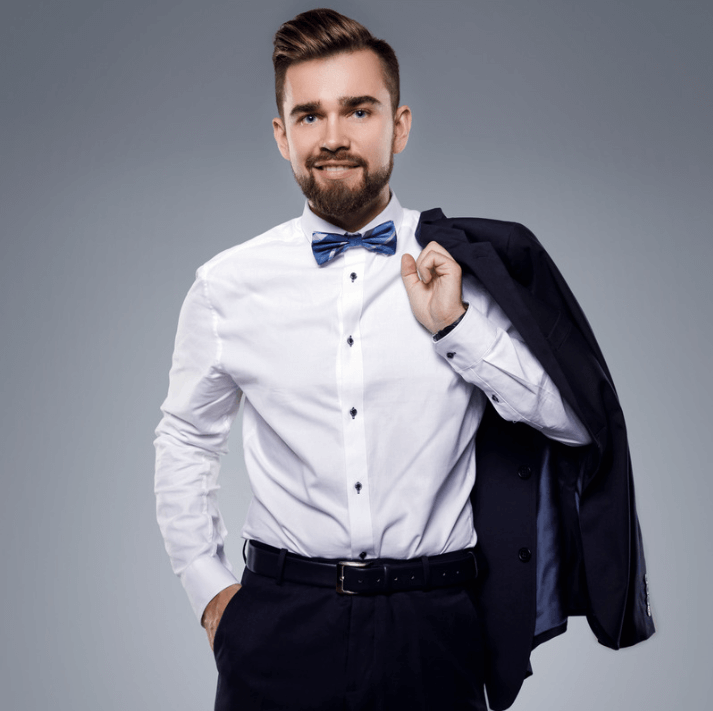 Muž v obleku s motýlikom a sako, usmievajúci sa na šedom pozadí.