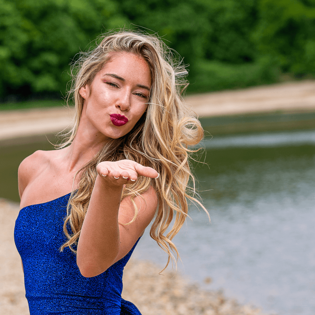 Žena v modrom šate s dlhými blond vlasmi, posielajúca bozk na pozadí prírody.